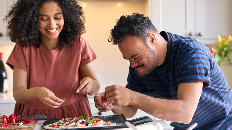 Two people smiling while preparing a pizza together in a kitchen